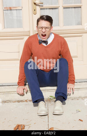 angry  man with open mouth with glasses and sweater sitting on steps in front of house and posing  while looking at camera, vint Stock Photo