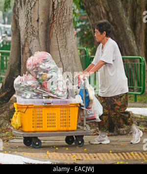 Asian woman on street in Singapore pushing trolley with plastic bins and bags filled with collected drink cans for recycling Stock Photo
