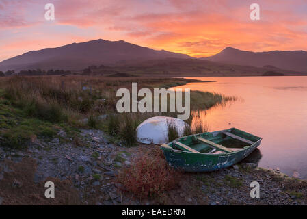 The sun rises behind mount Snowdon creating a beautiful orange sky which is reflected in the water of Llyn y Gader. Stock Photo