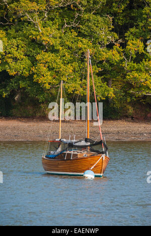 A varnished wooden sailing boat moored in the River Dart with a background of Autumn trees.