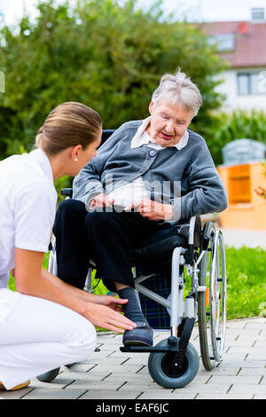 Nurse pushing senior woman in wheelchair on walk thru garden in summer Stock Photo