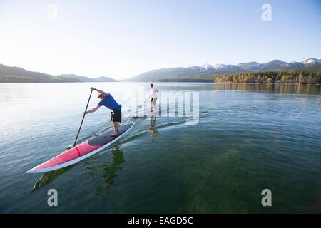 A fit male and female stand up paddle board (SUP) at sunset on Whitefish Lake in Whitefish, Montana. Stock Photo