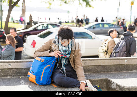 Young asian man wearing headphones watching a video on his mobile phone while sitting on the street in Seattle Stock Photo