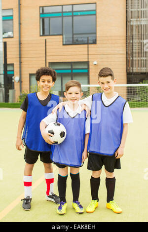 Portrait of three smiling boys wearing soccer uniforms and holding soccer ball in front of school Stock Photo