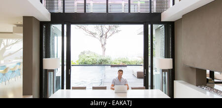 Young man using laptop in modern dining room with patio doors and swimming pool in background Stock Photo
