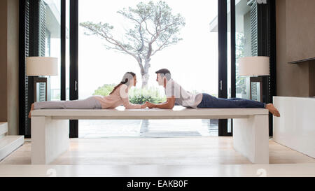 Couple lying on table face to face and holding hands, tree seen through patio door in background Stock Photo