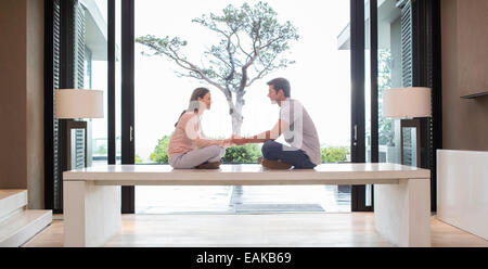 Couple sitting on table face to face and holding hands, tree seen through patio door in background Stock Photo