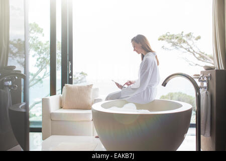 Woman wearing white bathrobe sitting on edge of modern bathtub and using digital tablet Stock Photo