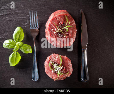 Pieces of red raw meat steaks with herbs, served on black stone surface. Shot from upper view. Stock Photo