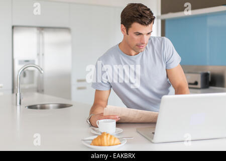 Man with newspaper using laptop at kitchen counter, coffee cup and croissant in foreground Stock Photo