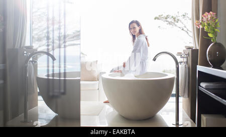 Portrait of smiling woman wearing white bathrobe, sitting on edge of bathtub in bathroom Stock Photo
