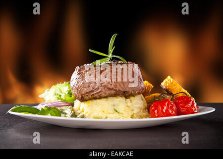 Piece of red meat steak with rosemary served on black stone surface. Blur fire flames on background Stock Photo