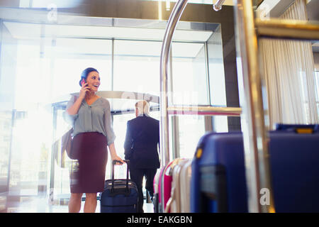 Smiling woman talking on phone in hotel lobby, luggage cart in foreground Stock Photo