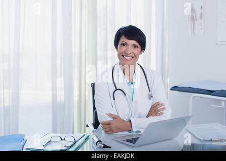 Portrait of smiling female doctor sitting with arms crossed at desk with laptop Stock Photo
