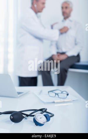 Blood pressure gauge, laptop and patient chart on desk in doctor's office, doctor examining patient in background Stock Photo
