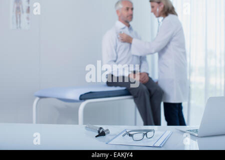 Female doctor examining patient in office, laptop, file, glasses and otoscope on desk in foreground Stock Photo
