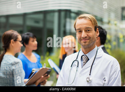 Portrait of smiling doctor wearing lab coat standing in front of hospital, women with clipboards in background Stock Photo