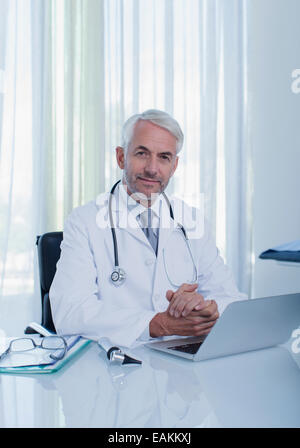 Portrait of smiling mature doctor sitting at desk with laptop in office Stock Photo