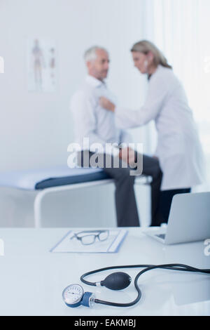 Blood pressure gauge, laptop, file and glasses on desk in doctor's office, female doctor examining patient in background Stock Photo