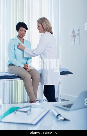 Female doctor examining her patient in office, laptop, otoscope, file and glasses on desk in foreground Stock Photo