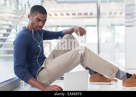 Portrait of businessman sitting on floor in modern office, using laptop and earphones Stock Photo
