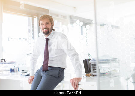 Portrait of smiling businessman wearing shirt and tie leaning on desk in office Stock Photo