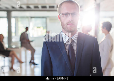 Portrait of serious businessman wearing glasses and suit standing in office, colleagues in background Stock Photo