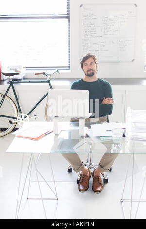Portrait of man sitting with arms crossed behind glass desk in modern office, bicycle and whiteboard in background Stock Photo