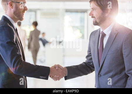 Businessmen wearing suits shaking hands in office, colleagues in background Stock Photo