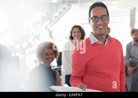 Portrait of smiling businessman wearing glasses and pink sweatshirt with team in background Stock Photo