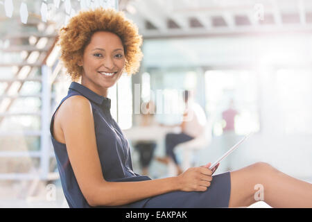Portrait of smiling woman using digital tablet in office corridor, colleagues in background Stock Photo