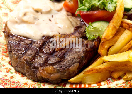 Closeup of grilled rib-eye beef steak served with mushroom sauce, salad and potato chips. Stock Photo