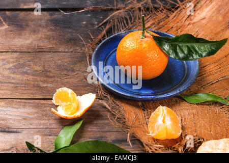 Whole and slice tangerines with leaves on blue ceramic plate over old wooden table. Stock Photo