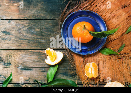 Whole and slice tangerines with leaves on blue ceramic plate over old wooden table. Top view. Stock Photo