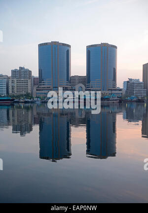 Two identical modern skyscrapers, smaller city buildings & pink clouds reflected in calm waters of Dubai Creek at dawn, UAE Stock Photo
