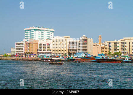 Colourful traditional wooden dhows & watertaxi (abra) on blue waters of Dubai Creek with buildings of adjacent city of Dubai rising into blue sky Stock Photo