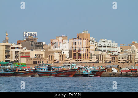 Colourful traditional wooden dhows moored at wharf on blue waters of Dubai Creek with buildings of adjacent city of Dubai rising into blue sky Stock Photo