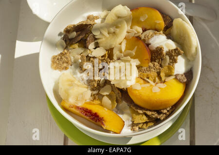 Breakfast in the sun, cereal bowl, peach and banana, yogurt on green plate and warm orange color. White on wood Stock Photo