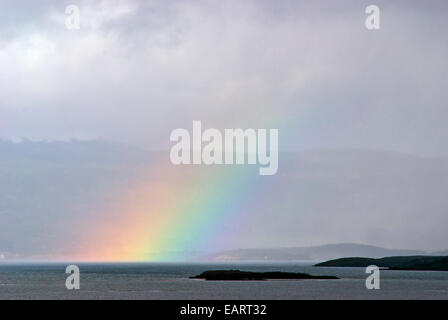 A rainbow rises from the ocean surface above a small rocky island. Stock Photo