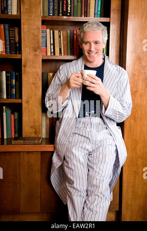 Middle-aged man leaning on bookshelf in pajamas with coffee Stock Photo
