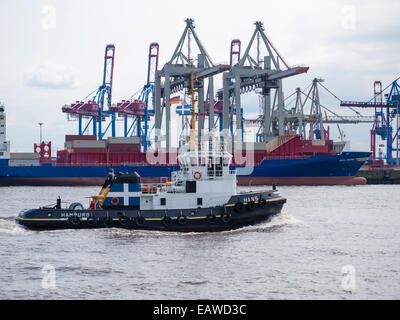 A tug boat is passing by cargo cranes of Hamburg's Burchardkai container terminal while a cargo ship is loaded in the back. Stock Photo