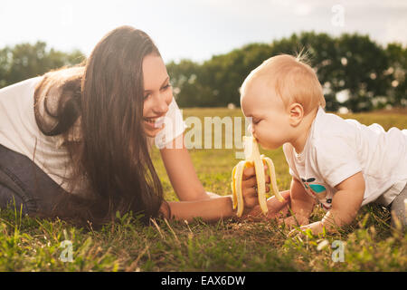 Young beautiful mother feeding her baby banana outdoors in sunlight Stock Photo