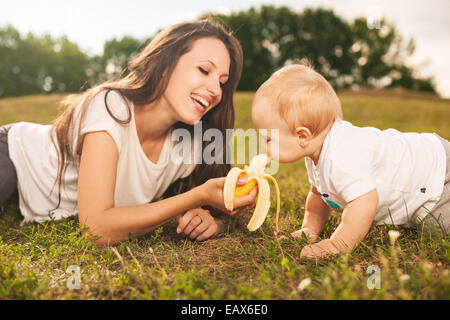 Young beautiful mother feeding her baby banana outdoors in sunlight Stock Photo