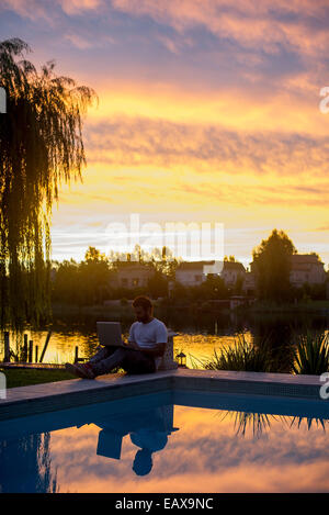 Man sitting next to pool using laptop computer Stock Photo
