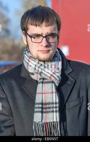 portrait of a serious young man in a black coat, scarf and glasses with a small beard and whiskers in autumn outdoors Stock Photo