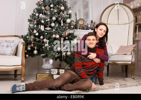 Pretty Woman Hugging Smiling Young Man While Resting on the Floor Carpet Near the Beautiful Christmas Tree and Large Elegant Cha Stock Photo