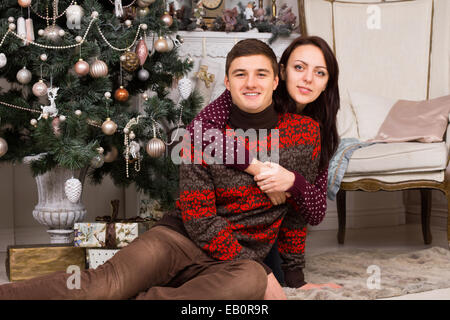 Close up Sweet Young White Couple Sitting on the Floor Carpet, Wearing Colored Trendy Winter Attire, Near Attractive Christmas T Stock Photo