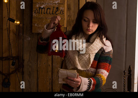 Pretty young woman in stylish colorful winter knitwear, emptying the mail box at night reading the information on a bundle of letters tied with string by lantern light. Stock Photo