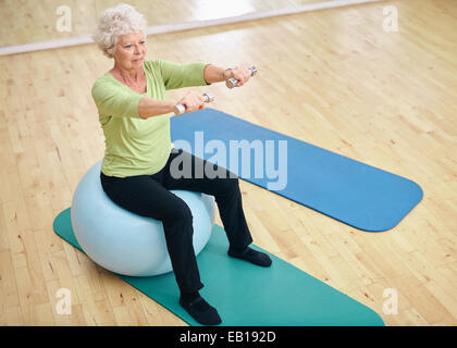 Senior female sitting on a fitness ball and lifting dumbbells. Old woman exercising with weights at gym.