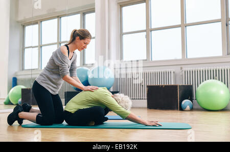 Female trainer helping senior woman to bend over. Old woman doing yoga on a exercise mat with physical therapist helping at gym. Stock Photo